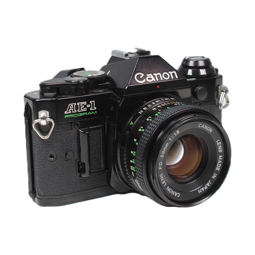 Front-left angle of black Canon AE1 Program 35mm film camera showing white logo, green aperture scale, and 50mm FD lens by ClubCamera.com