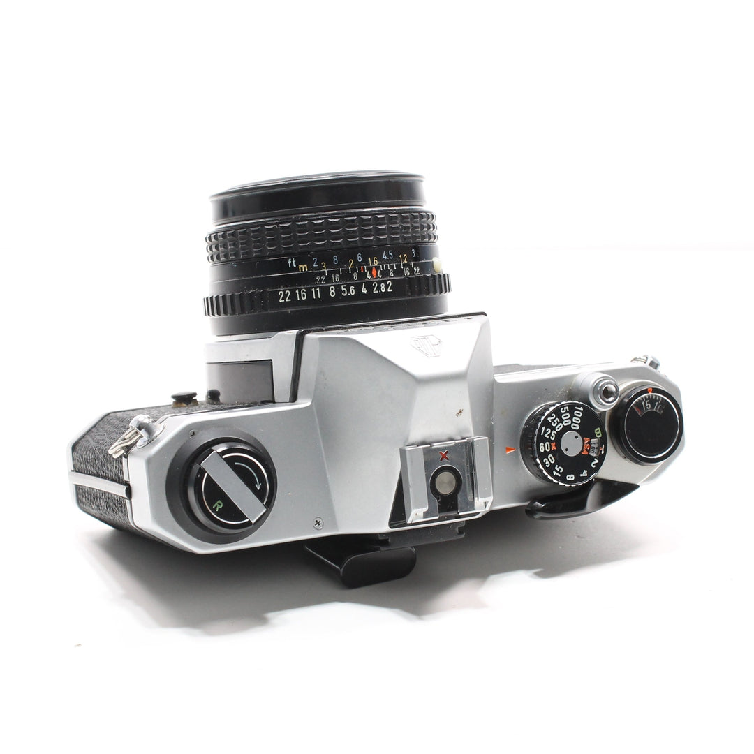 Top angled shot of silver Pentax K1000 35mm SLR kit showing shutter speed dial, aperture ring, and 50mm lens focus scale