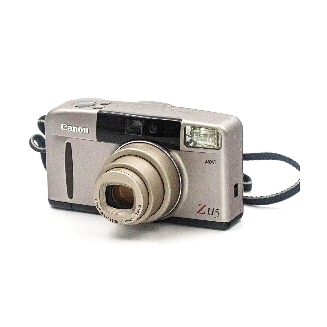Silver Canon Z115 compact film camera front three-quarter view showing gold zoom lens and ISYAF branding from ClubCamera.com